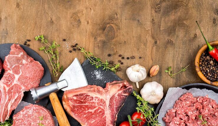 Carnes frescas y especias sobre una mesa de madera, incluyendo cortes de carne, carne molida, ajo, tomates cherry y chiles, con un hacha de cocina y ramitas de tomillo. Ideal para recetas de carne.
