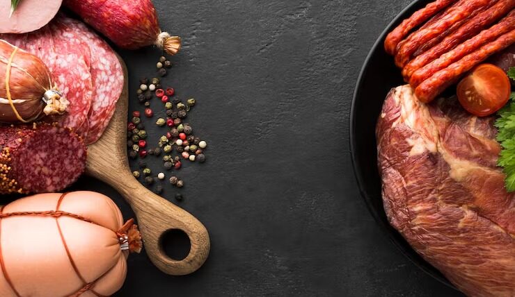 Selección de embutidos y carnes curadas sobre una superficie oscura, incluyendo salchichas, chorizo, y un trozo de carne fresca, acompañados de especias y hierbas.